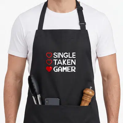 Gaming valentine's day apron - SINGLE TAKEN GAMER text with red outline and pixel hearts stacked vertically — shown on a black cotton canvas apron against a white background