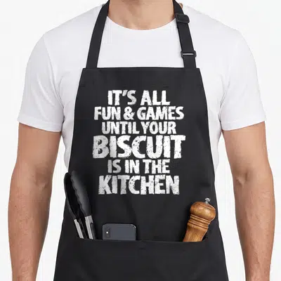 Shuffleboard joke apron - White stacked text reading IT'S ALL FUN & GAMES UNTIL YOUR BISCUIT IS IN THE KITCHEN — shown on a black cotton canvas apron against a white background