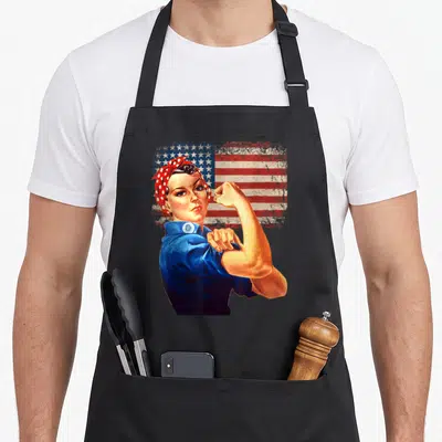 Rosie american flag apron - Woman flexing bicep in blue shirt against distressed red white and blue flag — shown on a black cotton canvas apron against a white background