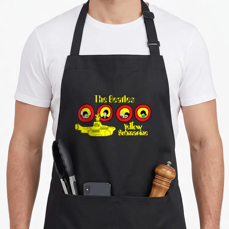 Cartoon yellow submarine apron - Yellow submarine graphic with four red circle portraits and Beatles text on black — shown on a black cotton canvas apron against a white background