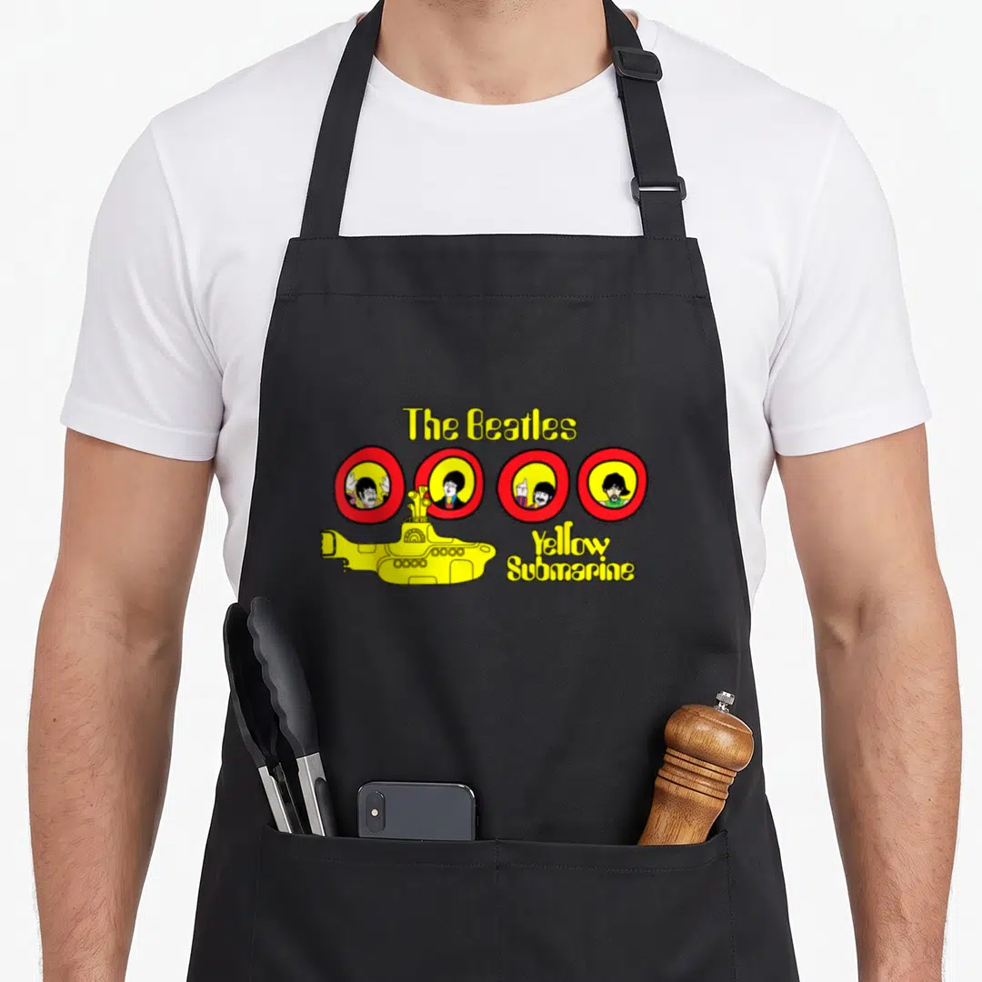 Cartoon yellow submarine apron - Yellow submarine graphic with four red circle portraits and Beatles text on black — shown on a black cotton canvas apron against a white background