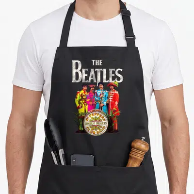 Sgt band apron - Beatles members in colorful uniforms behind a drum with printed band name text — shown on a black cotton canvas apron against a white background