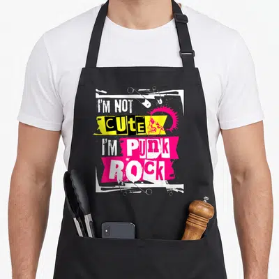 Funny rocker punk rock apron - Pink mohawk skull with safety pins and stacked text I'M NOT CUTE I'M PUNK on yellow — shown on a black cotton canvas apron against a white background