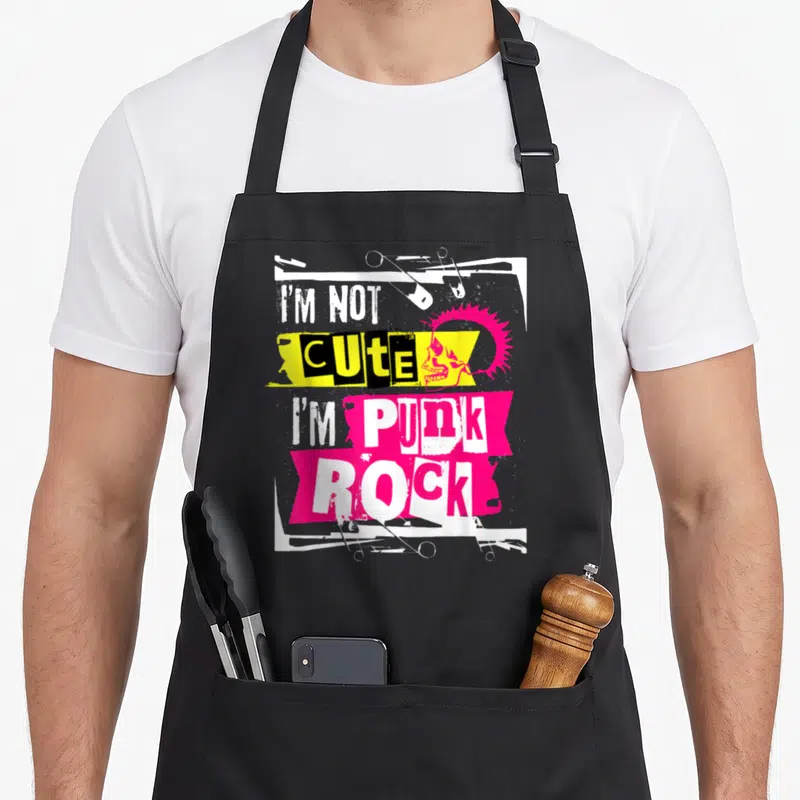 Funny rocker punk rock apron - Pink mohawk skull with safety pins and stacked text I'M NOT CUTE I'M PUNK on yellow — shown on a black cotton canvas apron against a white background