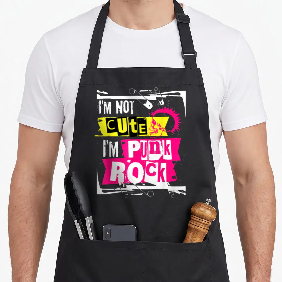 Funny rocker punk rock apron - Pink mohawk skull with safety pins and stacked text I'M NOT CUTE I'M PUNK on yellow — shown on a black cotton canvas apron against a white background