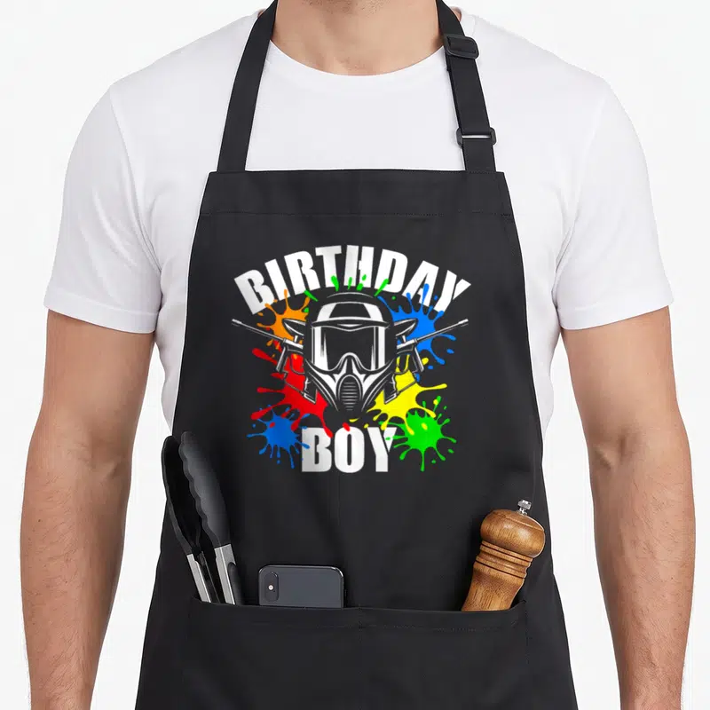 Paintball theme birthday apron - Cartoon paintball mask with crossed markers and white BIRTHDAY BOY text surrounded — shown on a black cotton canvas apron against a white background