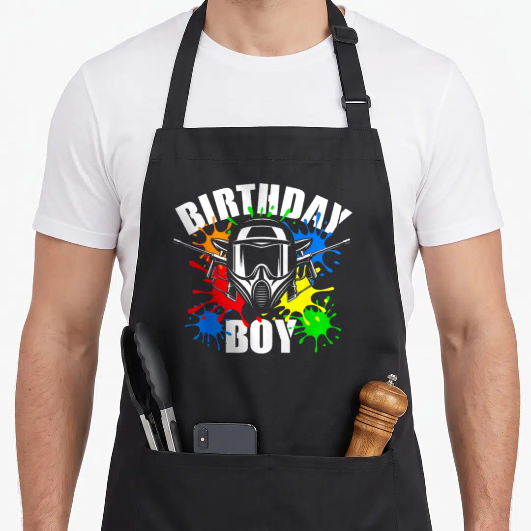 Paintball theme birthday apron - Cartoon paintball mask with crossed markers and white BIRTHDAY BOY text surrounded — shown on a black cotton canvas apron against a white background