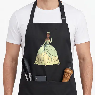 Funny frog princess apron - Cartoon Tiana in light green and yellow ballgown waving with brown skin tone — shown on a black cotton canvas apron against a white background