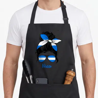 Nicaraguense nicaraguan girl apron - Stylized woman silhouette with bun and sunglasses in flag pattern, text Nica below — shown on a black cotton canvas apron against a white background