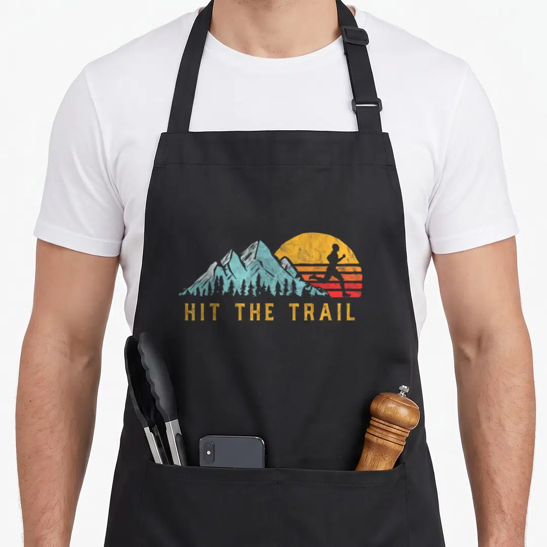 Hit runner apron - Retro runner silhouette against orange sun and teal mountains with HIT THE TRAIL text — shown on a black cotton canvas apron against a white background