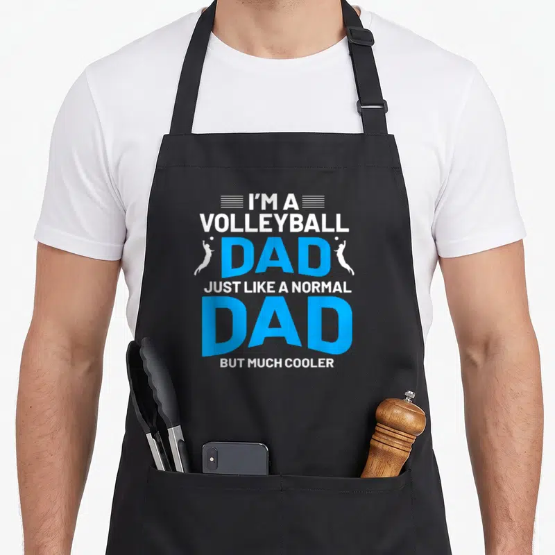 Cute volleyball dad apron - Stacked text I'M A VOLLEYBALL DAD with two white jumping player silhouettes flanking — shown on a black cotton canvas apron against a white background