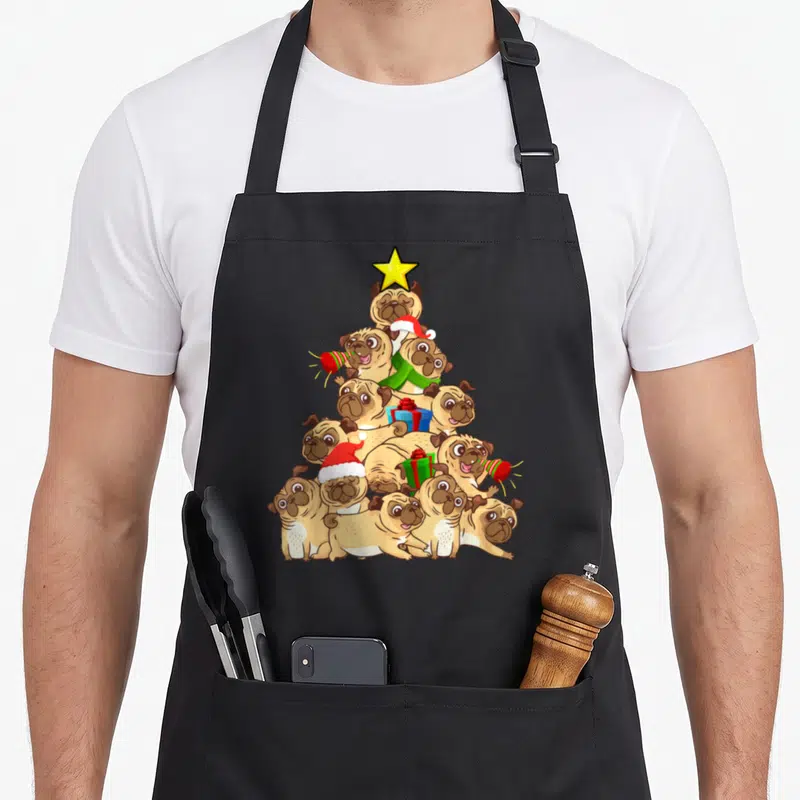 Sweater christmas tree apron - Cartoon tan pugs stacked in a Christmas tree pyramid with yellow star and red green — shown on a black cotton canvas apron against a white background