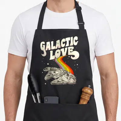 Galactic falcon apron - Retro Millennium Falcon with rainbow trail under GALACTIC LOVE text — shown on a black cotton canvas apron against a white background