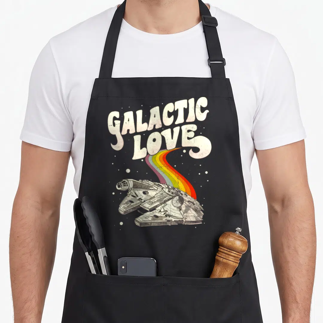 Galactic falcon apron - Retro Millennium Falcon with rainbow trail under GALACTIC LOVE text — shown on a black cotton canvas apron against a white background