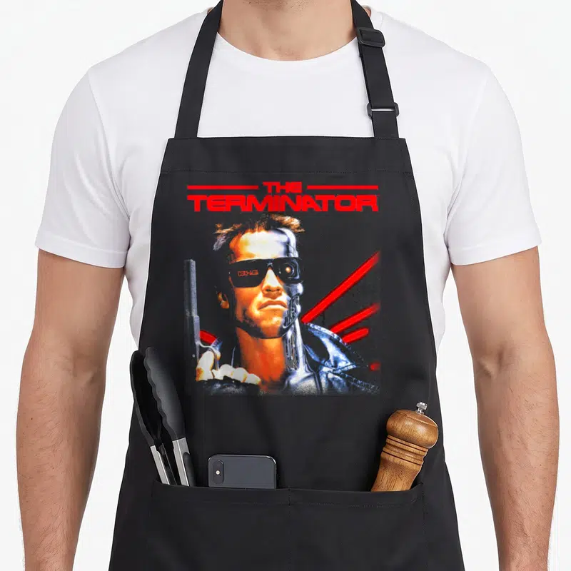 Terminator retro futurism apron - Red THE TERMINATOR text above a half-flesh half-metal face with red diagonal lines — shown on a black cotton canvas apron against a white background