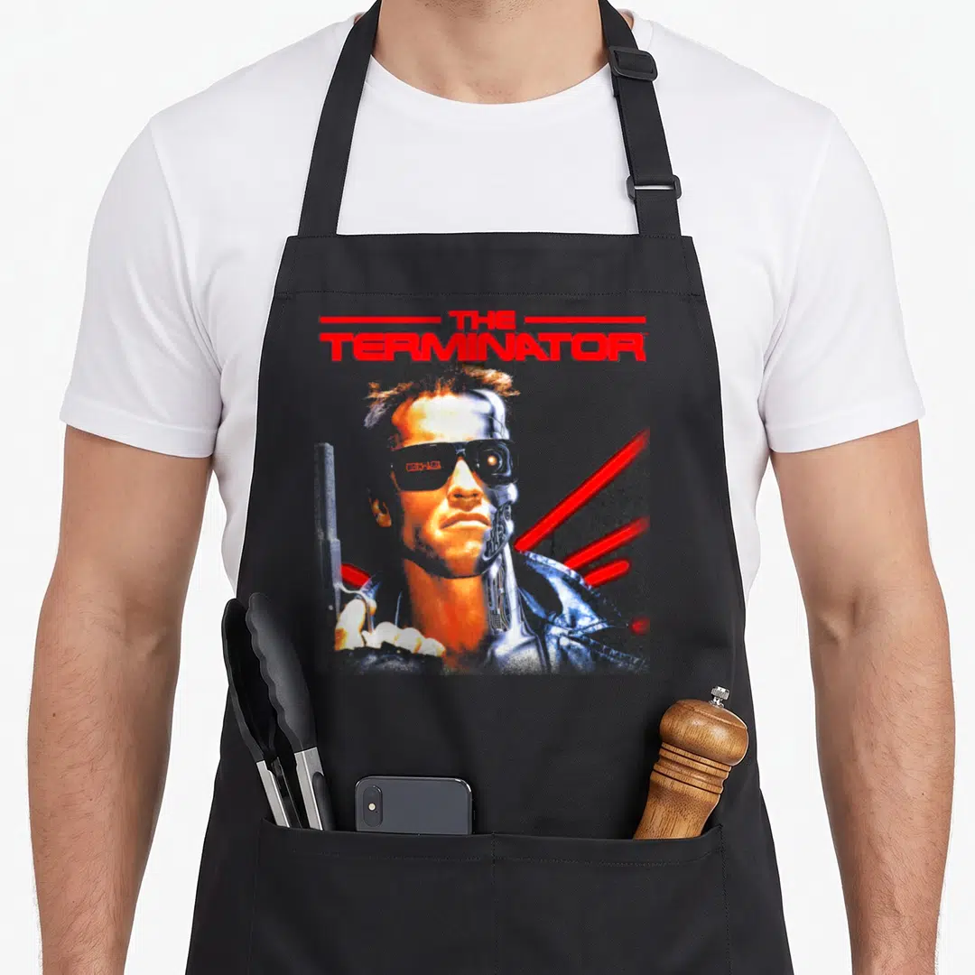 Terminator retro futurism apron - Red THE TERMINATOR text above a half-flesh half-metal face with red diagonal lines — shown on a black cotton canvas apron against a white background