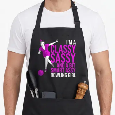 Assy smart apron - Cartoon girl in magenta outfit bowling with stacked white and pink text — shown on a black cotton canvas apron against a white background