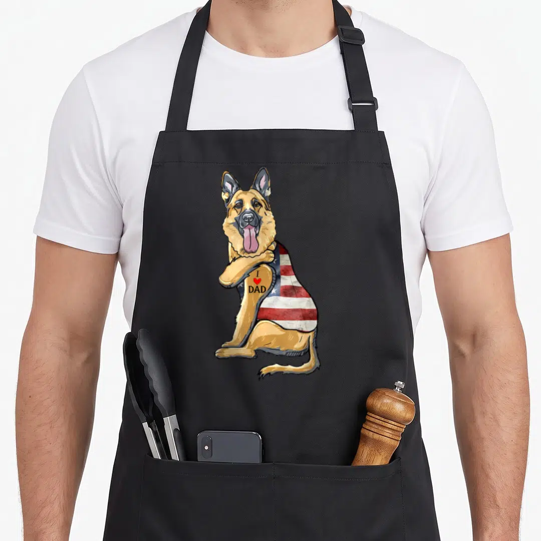 German tattoo aprons - Cartoon German Shepherd with USA flag and I Love Dad tattoo text — shown on a black cotton canvas apron against a white background