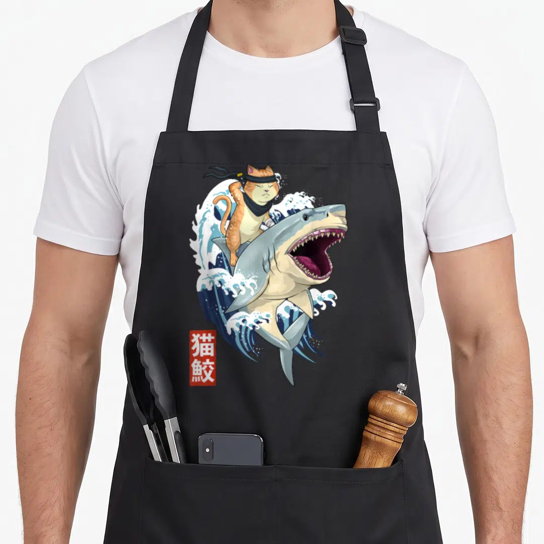 Kanagawa cat apron - Orange cat on leaping shark with blue waves and red kanji seal — shown on a black cotton canvas apron against a white background