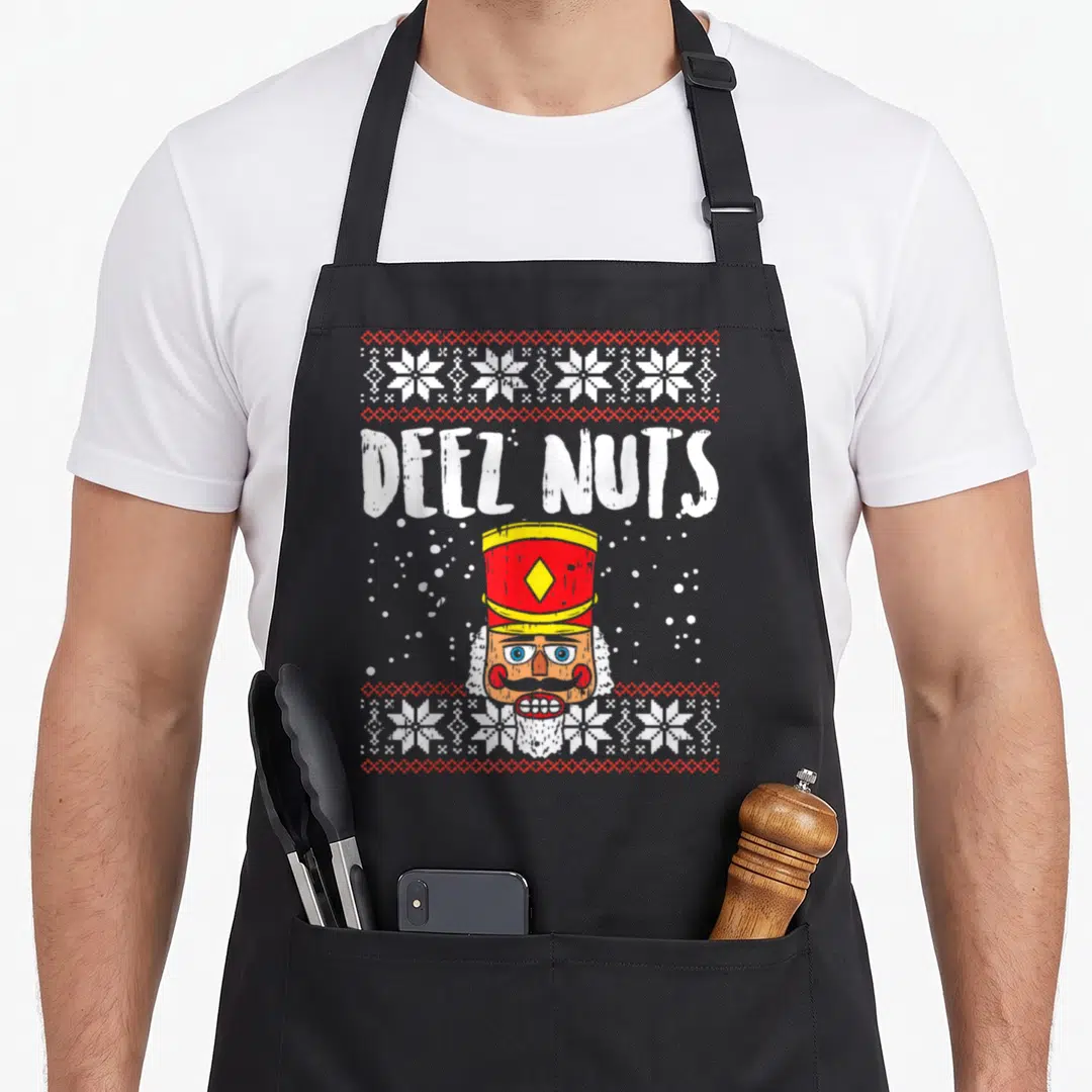Deez ugly christmas sweater apron - Deel Nutz text above a cartoon nutcracker head framed by snowflake bands and dots — shown on a black cotton canvas apron against a white background