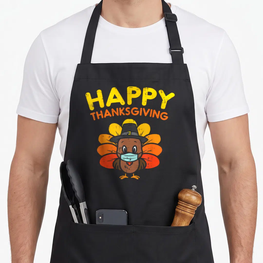 Mask turkey apron - Cartoon turkey in pilgrim hat and blue surgical mask under stacked HAPPY THANKSGIVING text — shown on a black cotton canvas apron against a white background