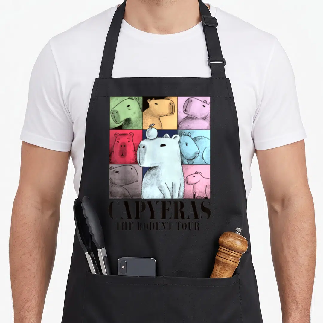 Capyeras animal apron - Cartoon capybara with apple on head below colorful grid of capybara heads and text reading — shown on a black cotton canvas apron against a white background
