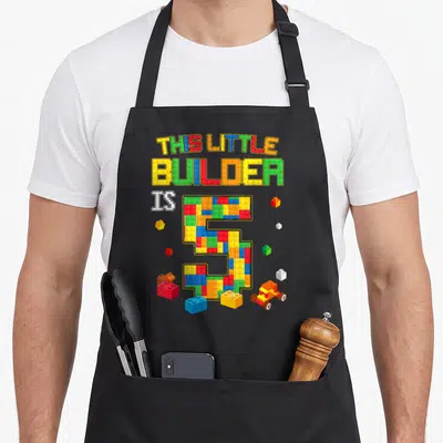Funny block building aprons - Number 5 made of yellow red blue and green toy bricks with scattered toys and text — shown on a black cotton canvas apron against a white background