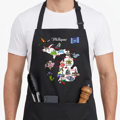 Illustrated michigan boy apron - White Michigan map with red city names and cartoon icons like deer, apple, and flag — shown on a black cotton canvas apron against a white background