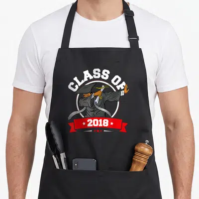 Graduation history class apron - Cartoon graduate dabbing holding diploma under arched CLASS OF text and red 2018 — shown on a black cotton canvas apron against a white background