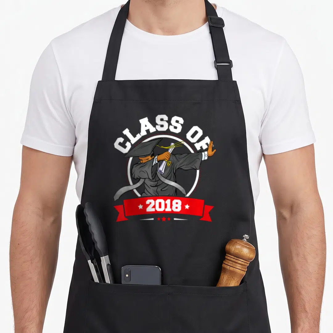 Graduation history class apron - Cartoon graduate dabbing holding diploma under arched CLASS OF text and red 2018 — shown on a black cotton canvas apron against a white background