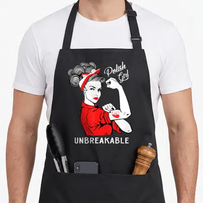 Unbreakable polish aprons - Cartoon woman flexing arm with Polish Girl script and Unbreakable text in red white black — shown on a black cotton canvas apron against a white background
