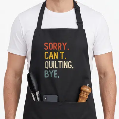 Sorry quilting apron - Stacked text SORRY CAN'T QUILTING BYE in red yellow off white and light blue typography — shown on a black cotton canvas apron against a white background