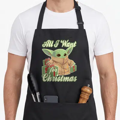General christmas apron - Green character in tan robe surrounded by red ribbon gift boxes under arched text — shown on a black cotton canvas apron against a white background