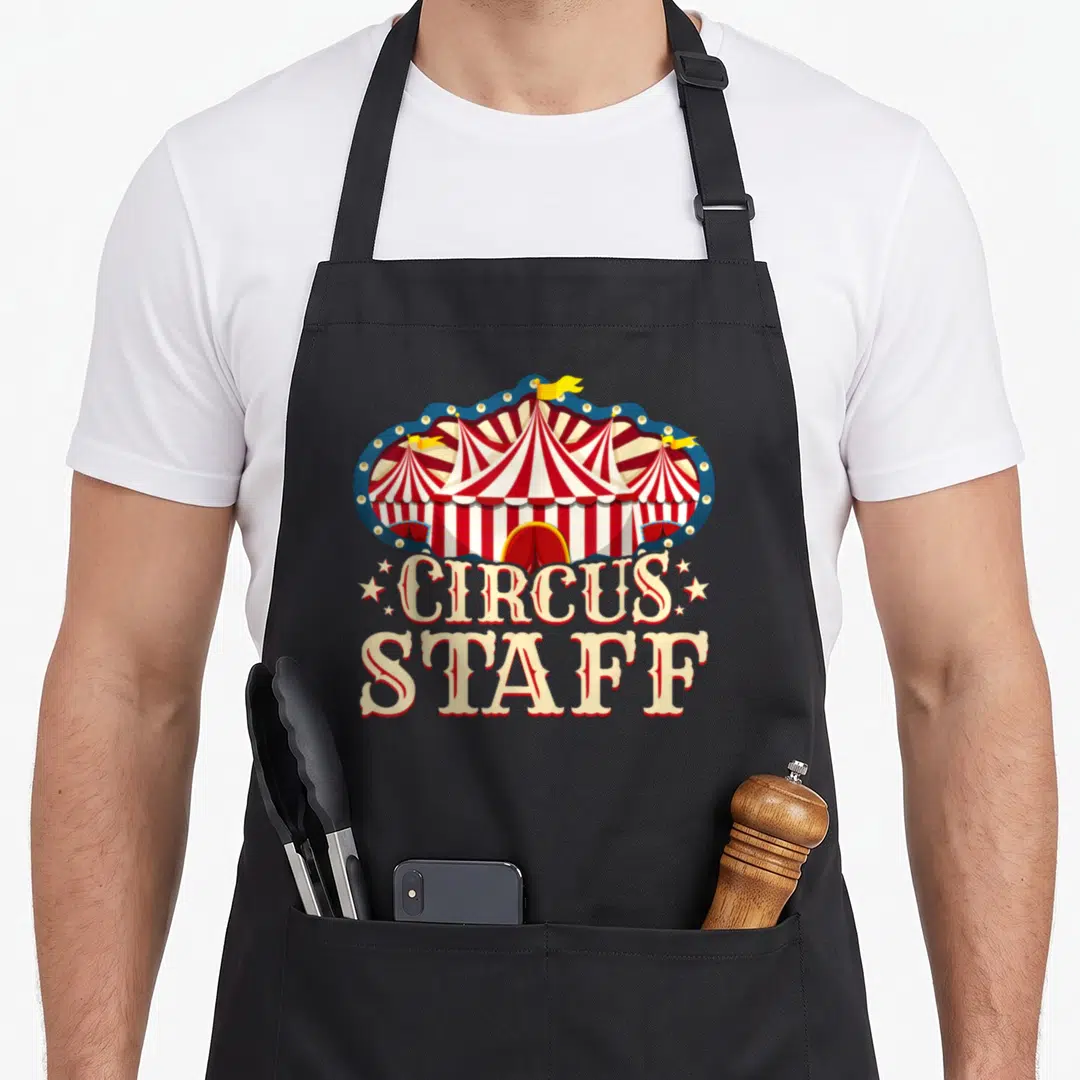 Circus staff aprons - Cartoon circus tent with CIRCUS STAFF text and scattered stars in red white yellow — shown on a black cotton canvas apron against a white background