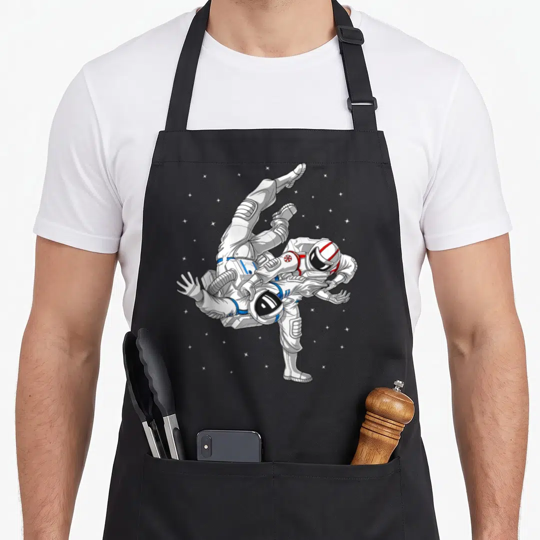 Astronaut brazilian jiu jitsu apron - Two cartoon astronauts grappling in space suits with red and blue details among — shown on a black cotton canvas apron against a white background