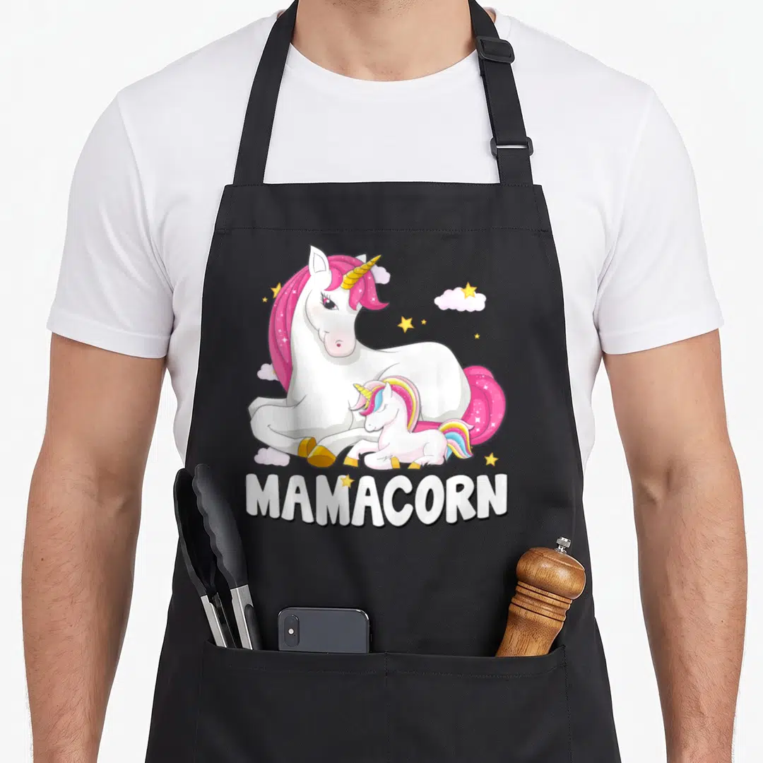 Baby mother's apron - White unicorn and baby with pink mane under MAMACORN text and yellow stars — shown on a black cotton canvas apron against a white background