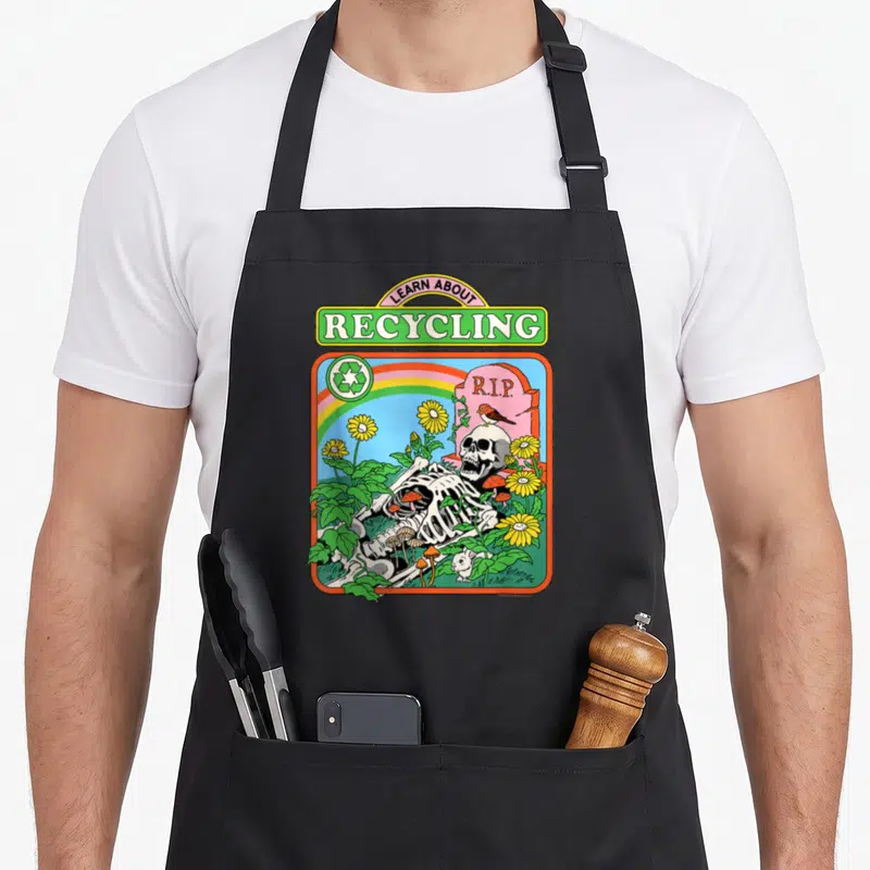 Steven skeleton apron - Cartoon skeleton lying near R.I.P. tombstone under rainbow with Learn About Recycling text — shown on a black cotton canvas apron against a white background