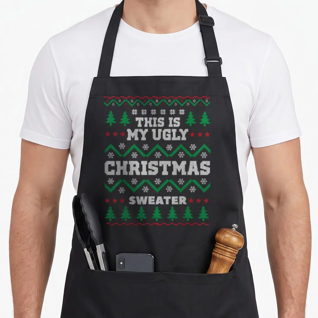 Mas ugly sweater apron - Stacked knit text reading THIS IS MY UGLY CHRISTMAS SWEATER with green trees red stars — shown on a black cotton canvas apron against a white background