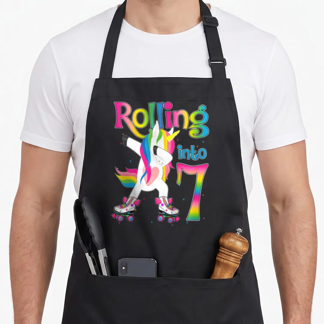 Rolling roll turning apron - Dabbing unicorn on roller skates with rainbow text Rolling into 7 and scattered stars — shown on a black cotton canvas apron against a white background