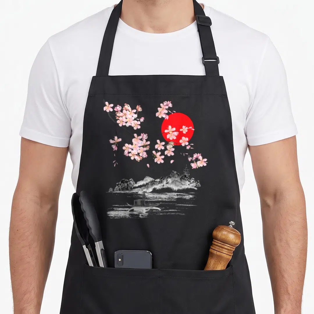 Blossom sakura cherry print apron - Pink cherry blossom branches over a red sun and a grey landscape with a boatman — shown on a black cotton canvas apron against a white background