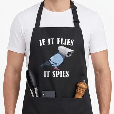 Spie meme apron - Pigeon with security camera head and text IF IT FLIES IT SPIES — shown on a black cotton canvas apron against a white background