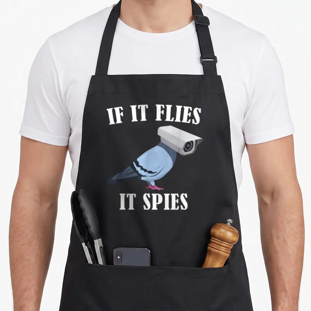 Spie meme apron - Pigeon with security camera head and text IF IT FLIES IT SPIES — shown on a black cotton canvas apron against a white background