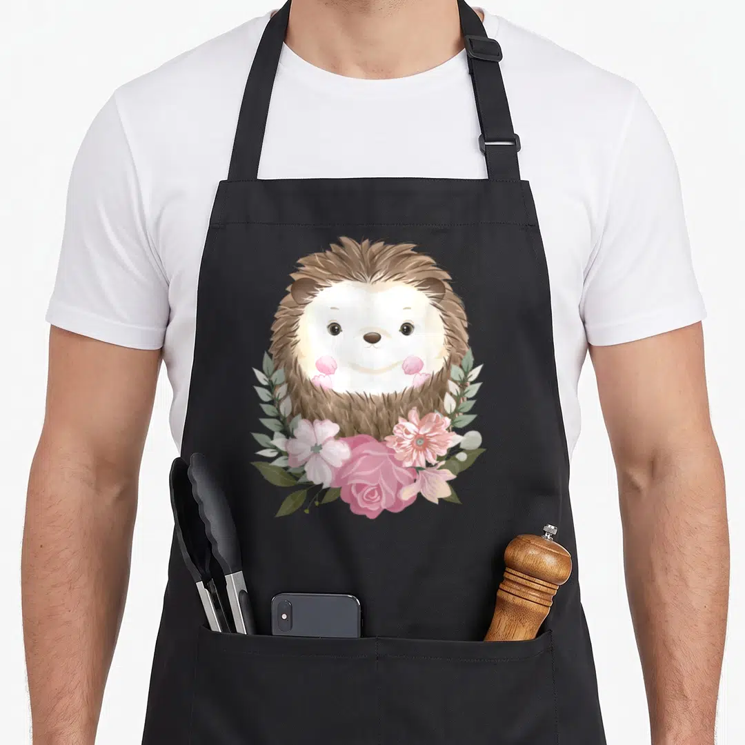 Hedge floral apron - Cartoon brown hedgehog face surrounded by pink flowers and green leaves — shown on a black cotton canvas apron against a white background