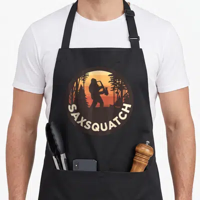 Sax bigfoot apron - Circular retro design of a sasquatch playing saxophone with curved text against a sunset forest — shown on a black cotton canvas apron against a white background
