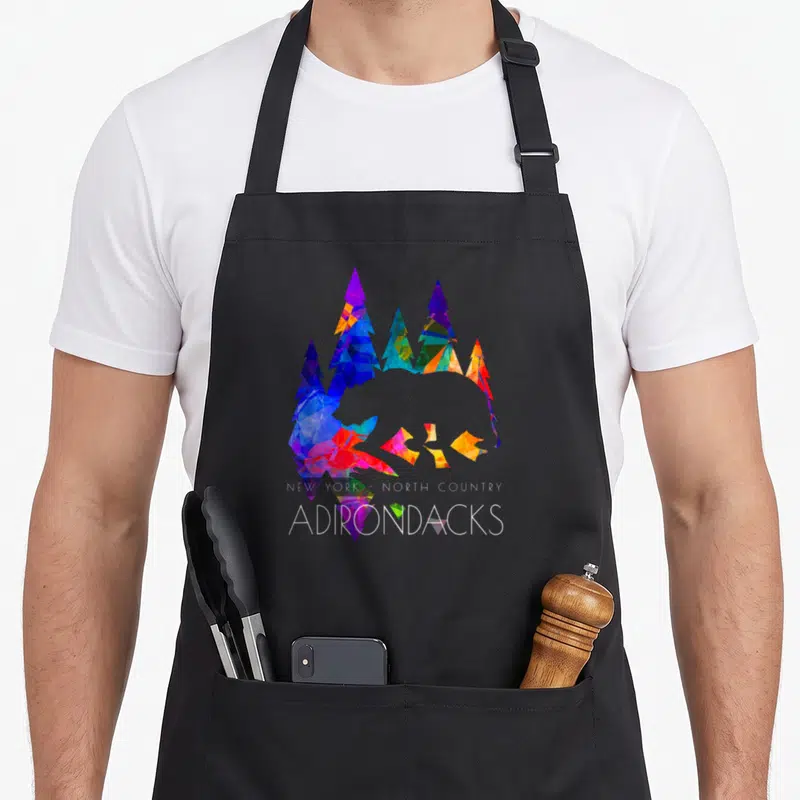 Bear new york apron - Geometric bear silhouette formed by blue purple red yellow and white pine trees with white text — shown on a black cotton canvas apron against a white background
