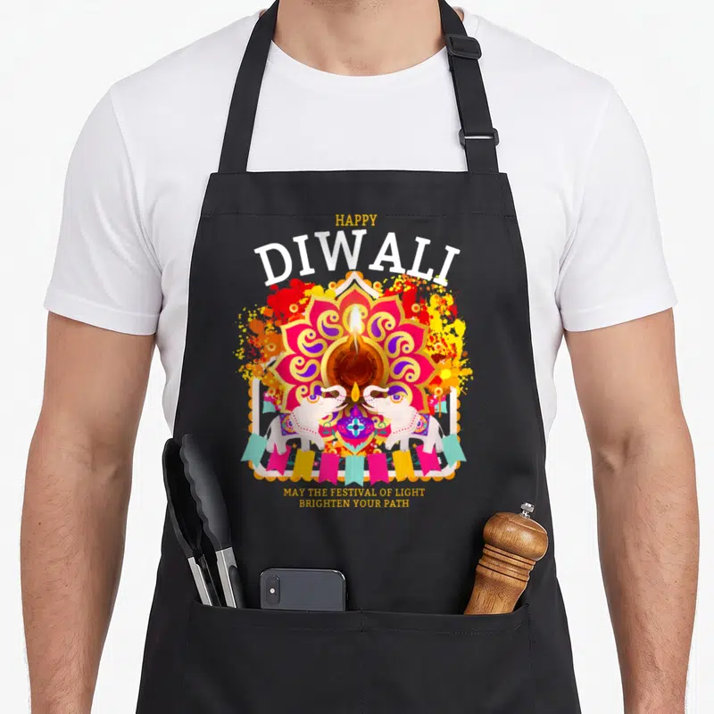 Outfit diwali apron - Lit diya in pink mandala with two white elephants and bunting flags under Happy Diwali text — shown on a black cotton canvas apron against a white background