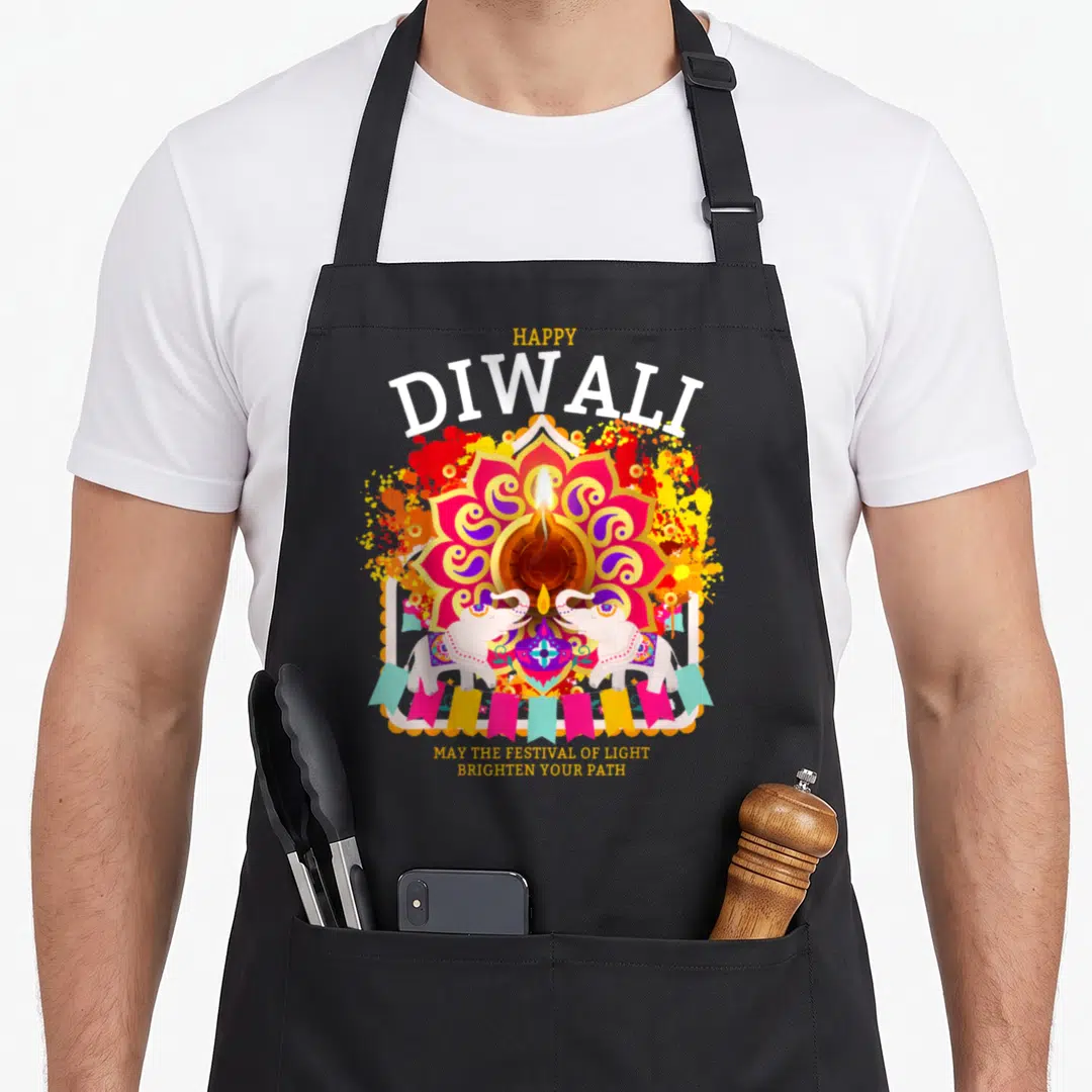 Outfit diwali apron - Lit diya in pink mandala with two white elephants and bunting flags under Happy Diwali text — shown on a black cotton canvas apron against a white background