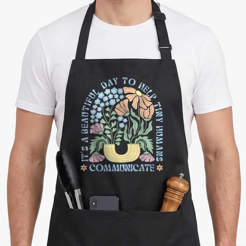 Pathologist pathology my therapy apron - Cartoon flowers in a yellow vase with blue text arching over and Communicate — shown on a black cotton canvas apron against a white background