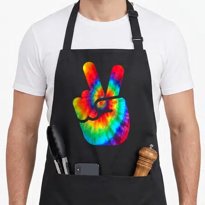 Peace tie-dye apron - Rainbow tie-dye hand making a peace sign with two extended fingers on a black background — shown on a black cotton canvas apron against a white background