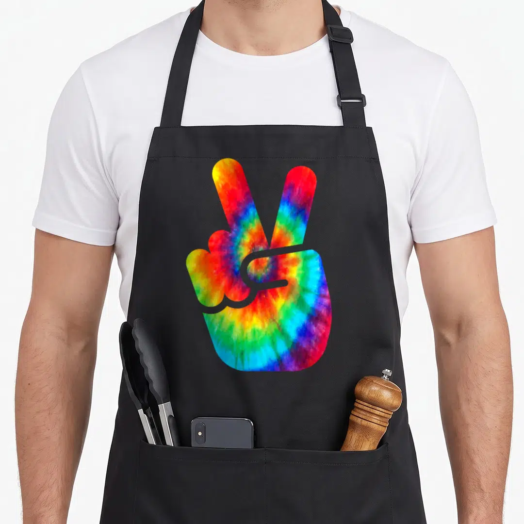 Peace tie-dye apron - Rainbow tie-dye hand making a peace sign with two extended fingers on a black background — shown on a black cotton canvas apron against a white background
